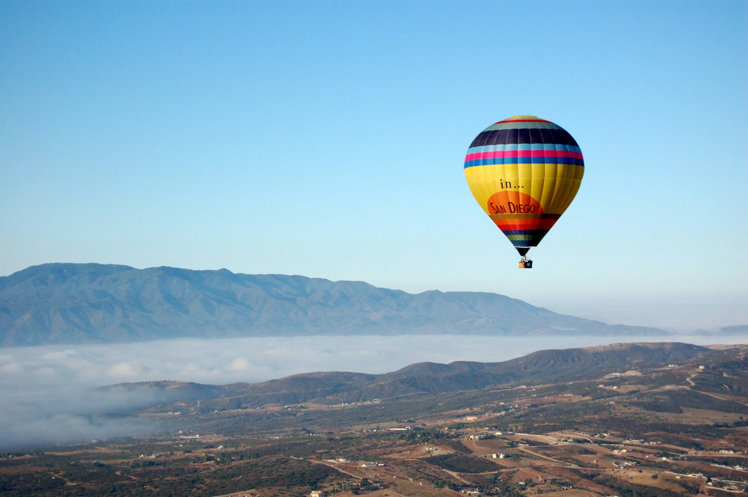 A colorful hot air balloon flying over mountainous terrain and a layer of clouds, with a clear blue sky in the background.