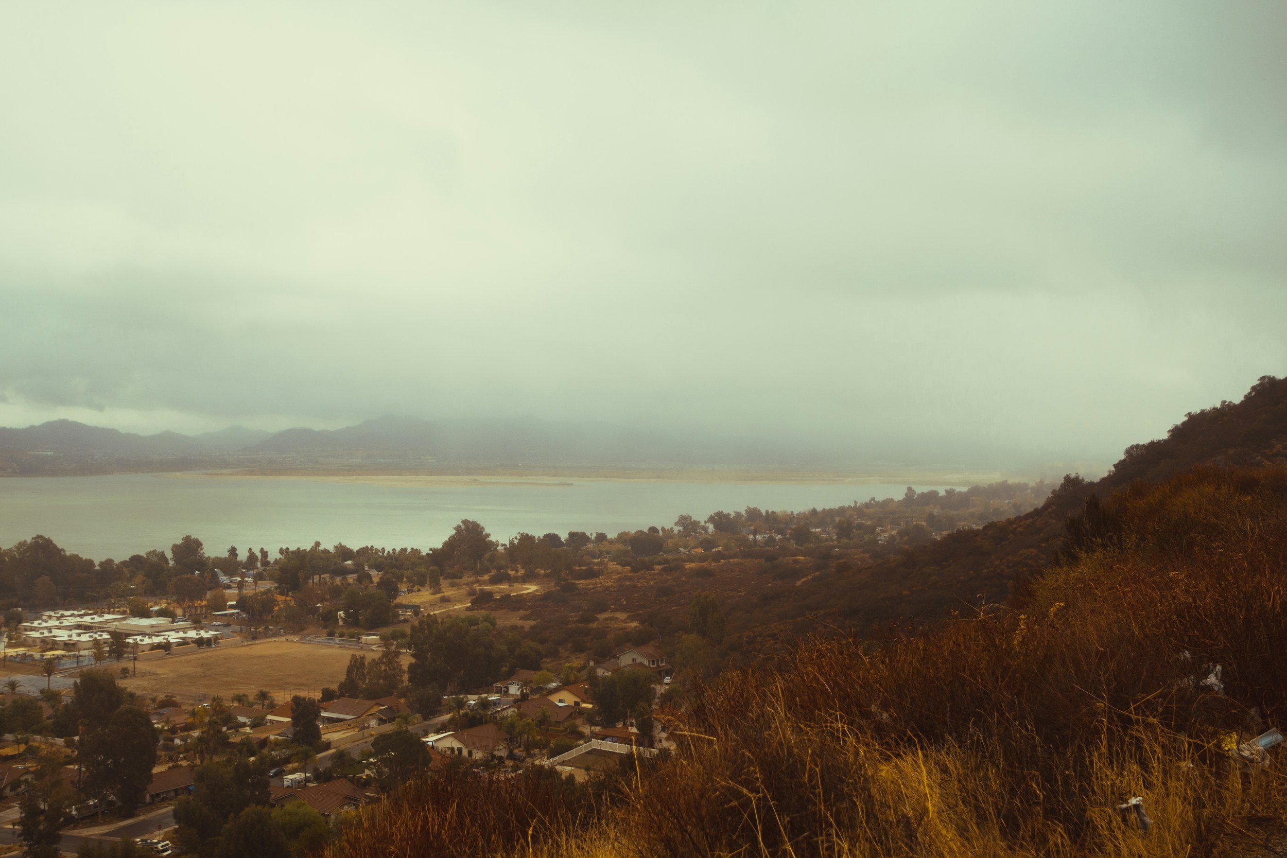 A scenic view of a residential area near a body of water with hilly terrain and a cloudy sky.