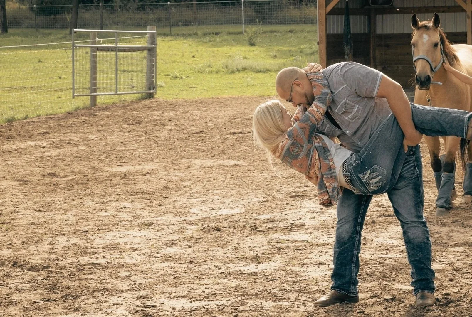 A man and a woman engaged in a playful wrestling or dancing moment in a dirt area at a farm or ranch, with a horse nearby and a wooden barn in the background.