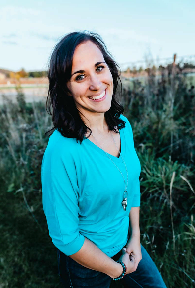 A woman with dark hair, wearing a bright blue top and a necklace, standing outdoors in a grassy area with a fence and buildings in the background, smiling at the camera.
