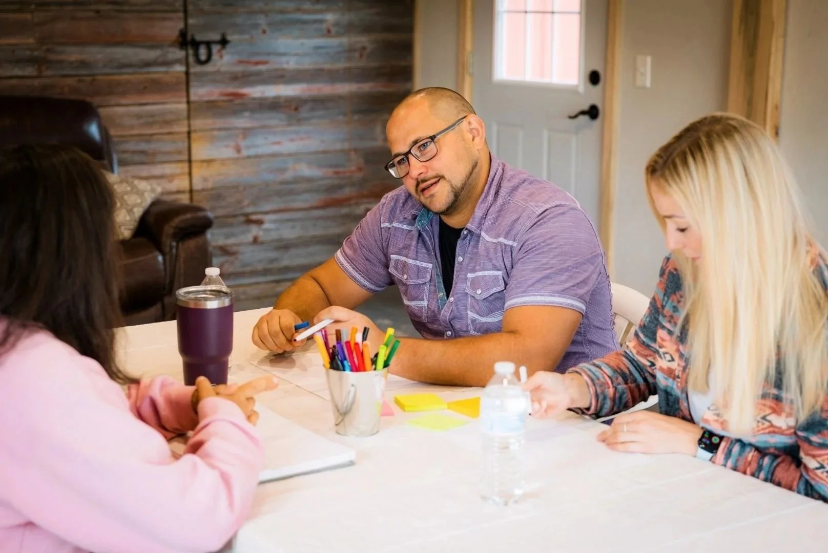 A man sitting at a table with two women, engaged in a discussion, with colorful markers, sticky notes, and water bottles on the table, in a room with wooden walls and a door behind them.