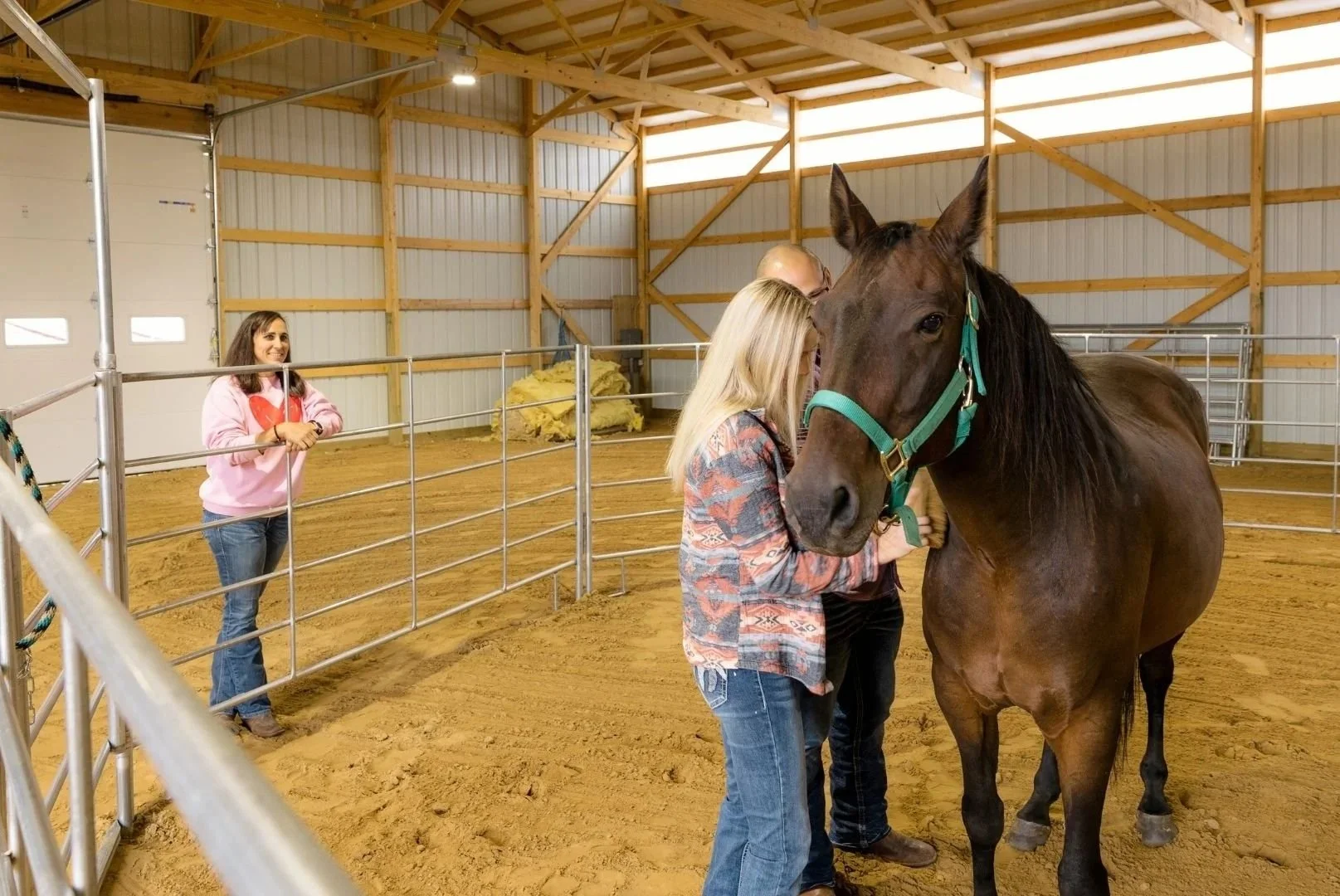 A woman with blonde hair and a patterned shirt stands next to a brown horse with a teal halter inside a wooden riding barn, while another woman in a pink sweatshirt leans against a metal railing watching them.
