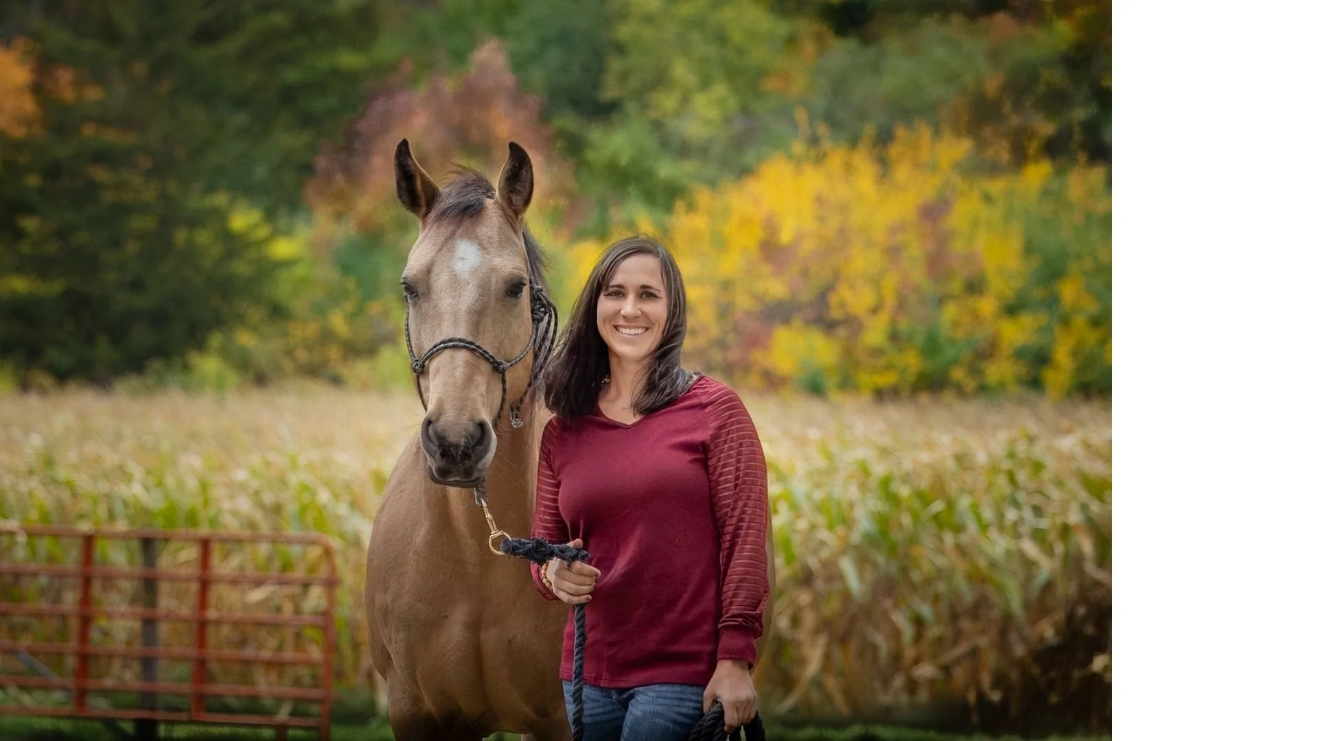 A woman with long dark hair wearing a red long sleeve shirt standing next to a light brown horse with a white star on its forehead, outdoors during fall with colorful autumn trees in the background.