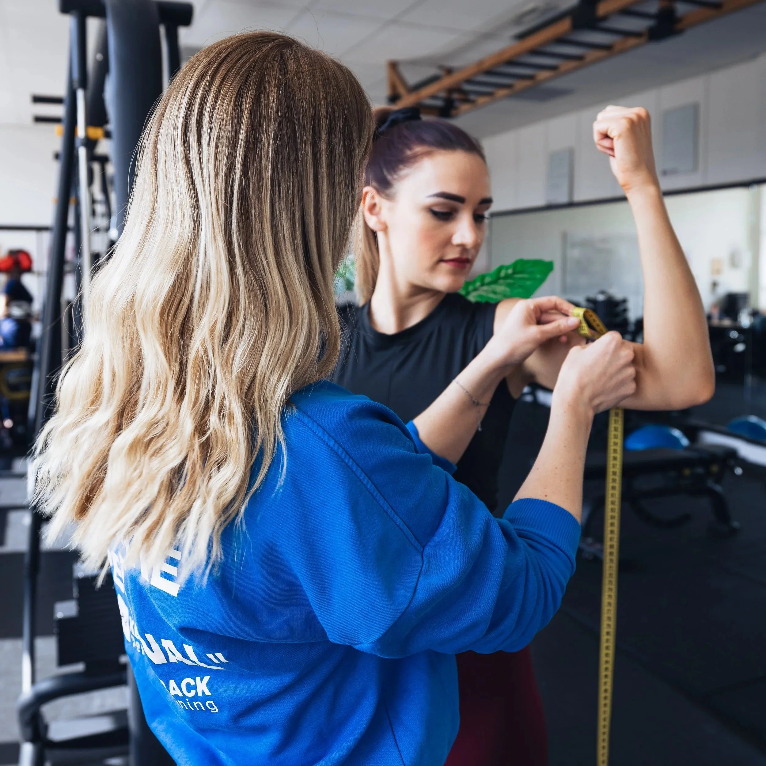 Twee vrouwen in een sportschool, een van hen meet haar arm met een meetlint, terwijl de ander haar begeleidt.