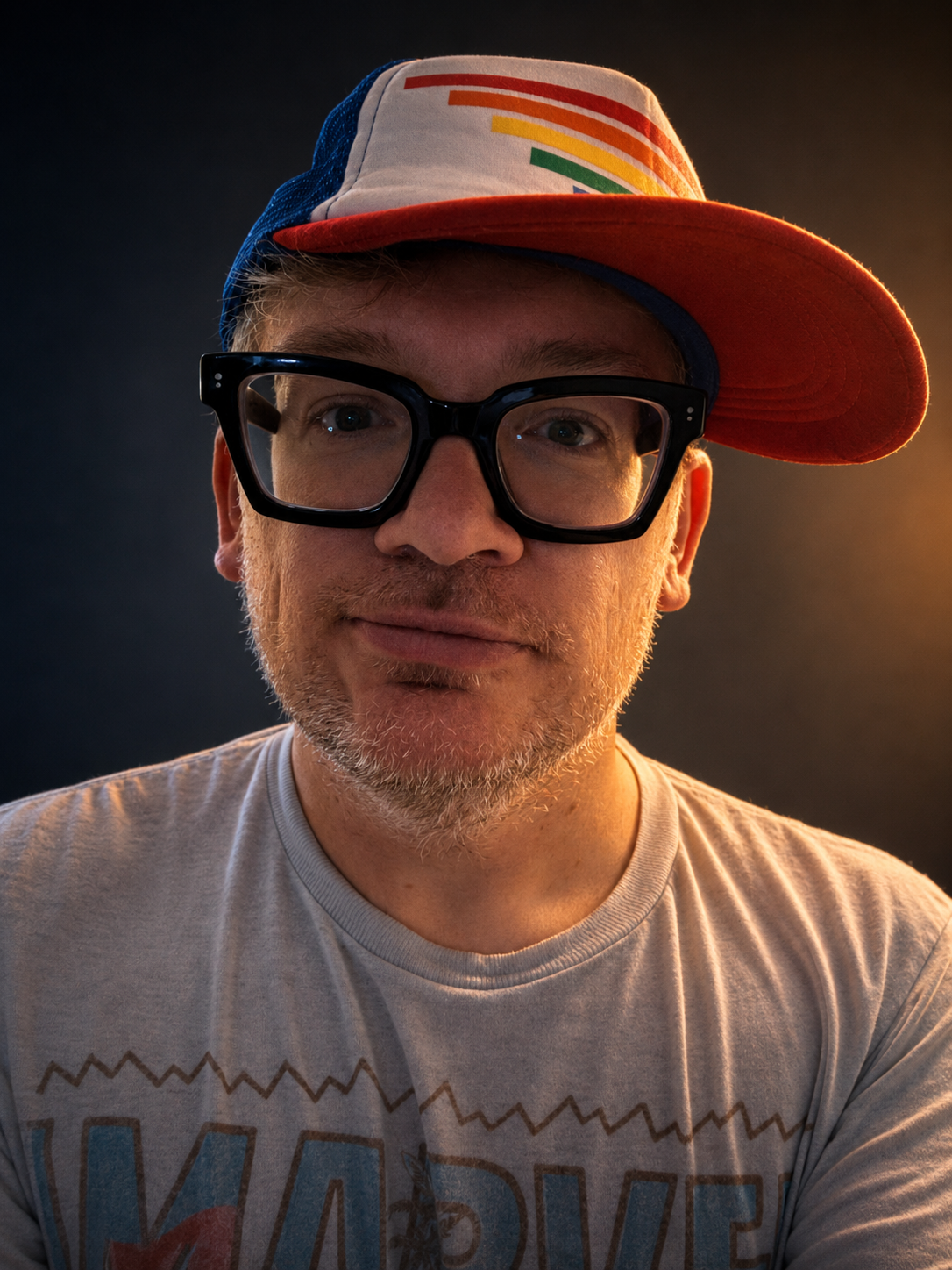 A man wearing a multicolored cap tilted to the side, large black glasses, and a t-shirt, with a dark background and warm lighting.