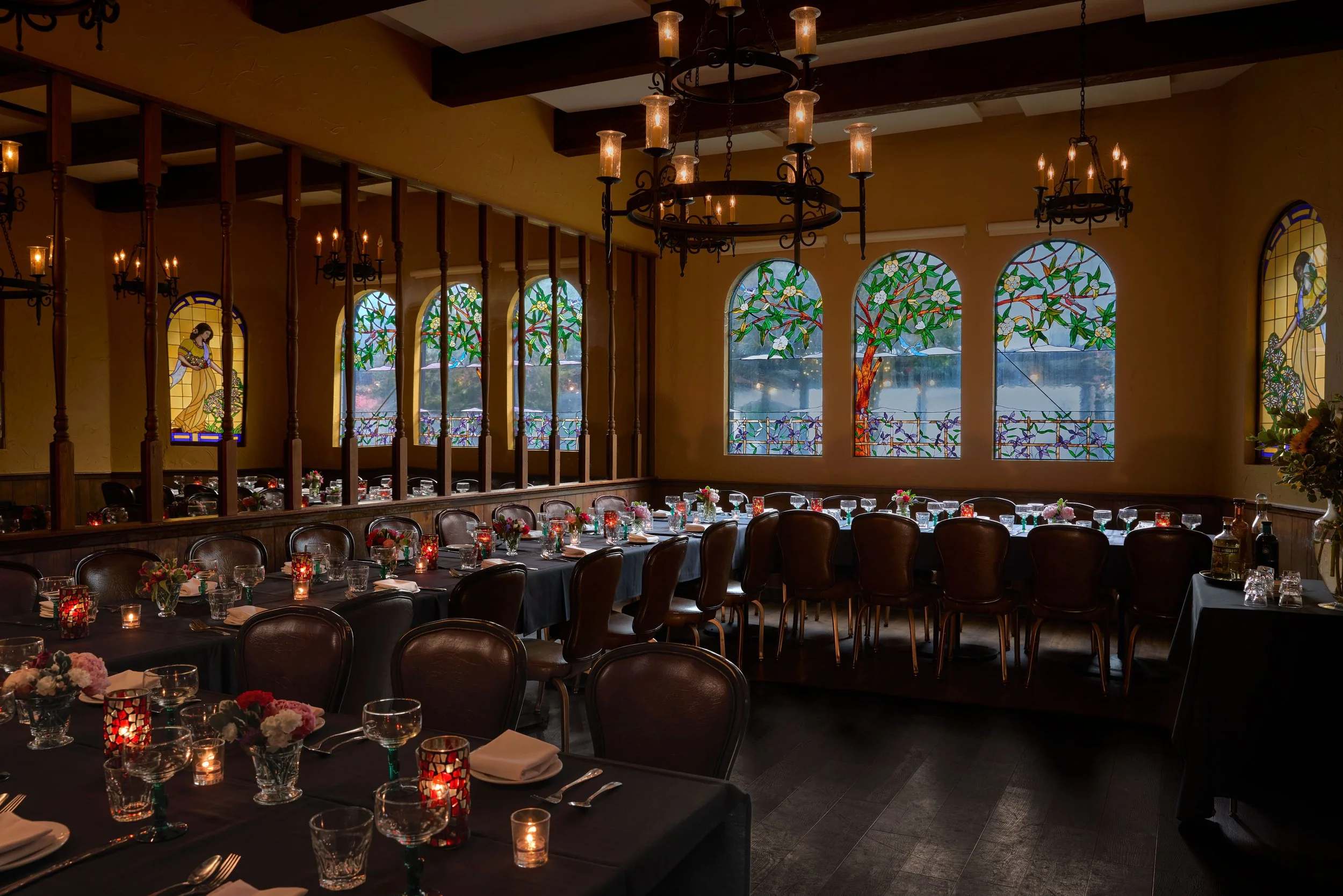 Elegant restaurant interior with decorated tables, floral centerpieces, wine glasses, and candles, featuring stained glass windows with floral and bird designs, wooden beams, and chandeliers.