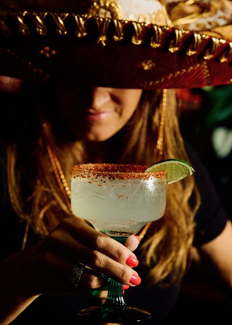 Woman wearing a large sombrero hat holding a margarita glass with a lime wedge and salted rim in a dimly lit setting.
