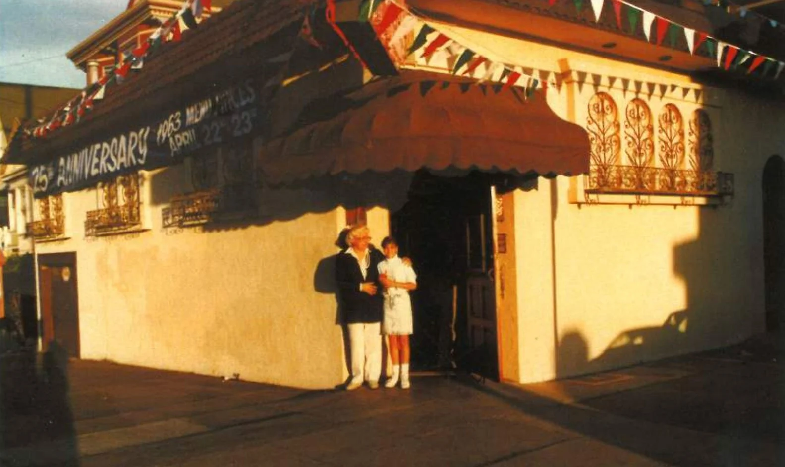 Celebrating the 25th anniversary of an event in April 1963 with two women standing in front of a building decorated with colorful flags and banners.