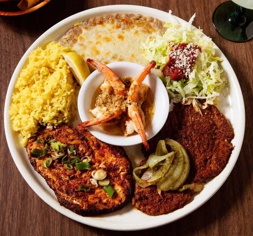 A plate of Southern comfort food including fried catfish topped with chopped green onions, yellow rice, baked macaroni and cheese, fried shrimp in the center, coleslaw with tomato and cheese, and a fried pork chop with sautéed green beans.