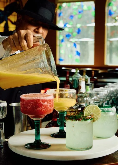 Bartender pouring yellow drink into colorful margarita glasses at a bar.