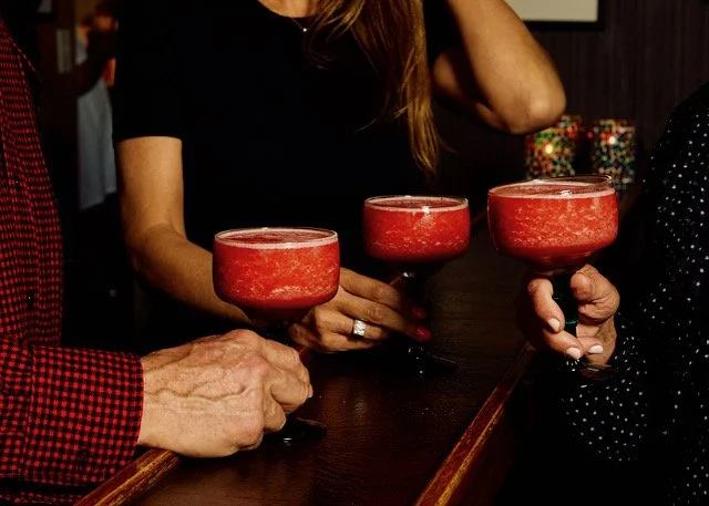 Three people holding red cocktails in a bar, with one person wearing a black shirt and the other in a polka dot shirt.