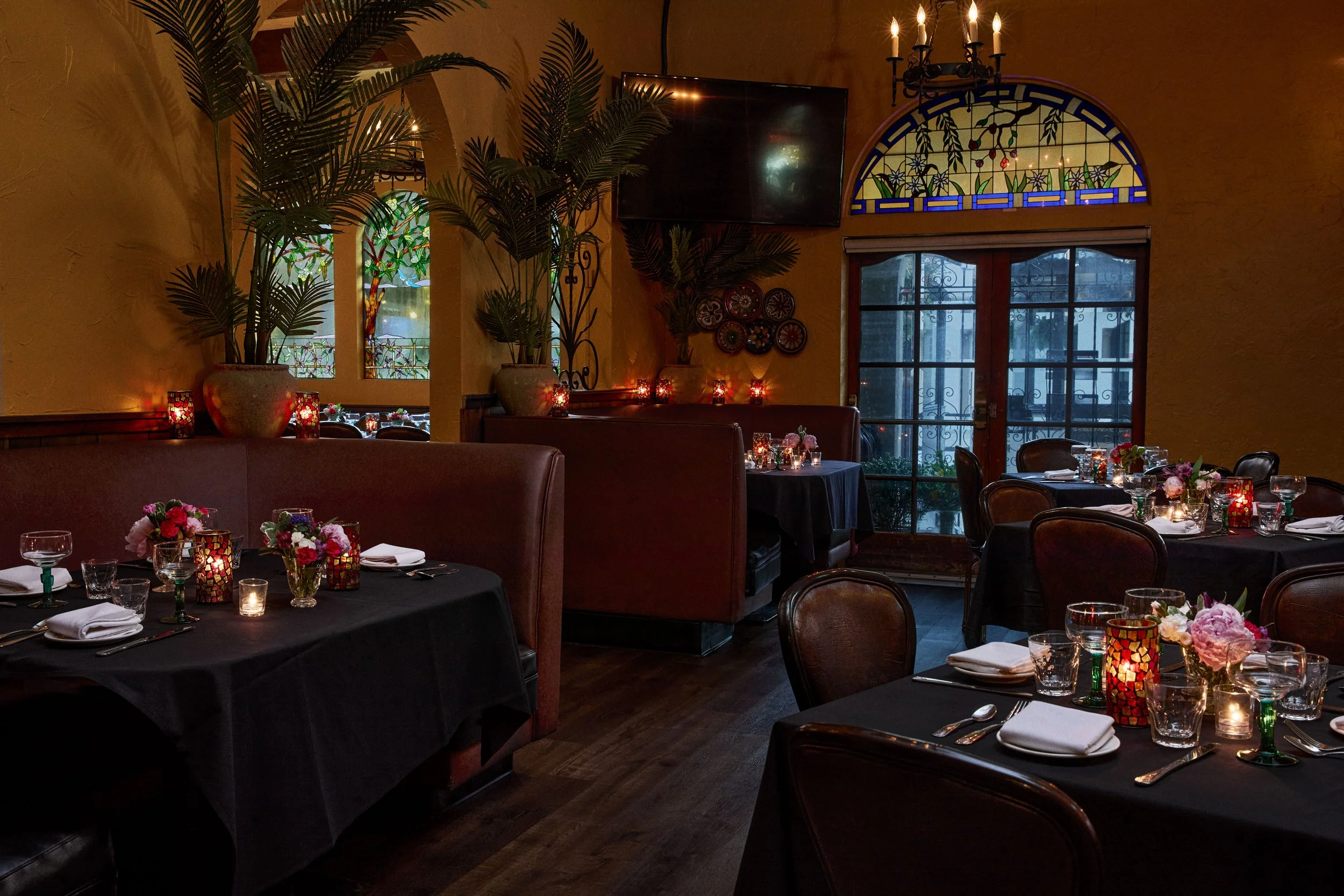 Interior of a restaurant with decorated tables, large potted plants, stained glass windows, and candlelit ambiance.