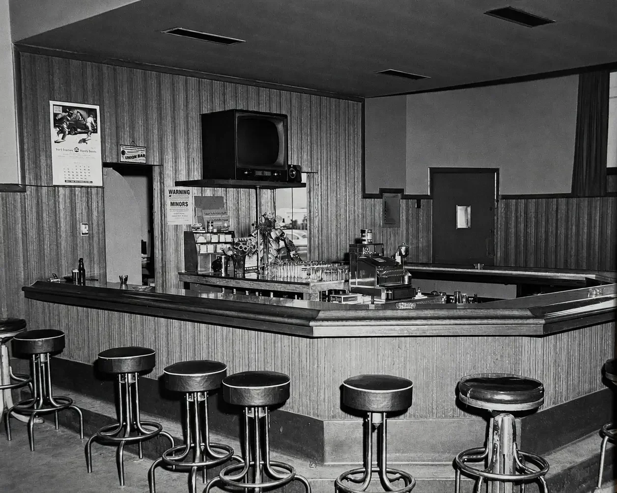 Empty vintage bar with a curved counter, barstools, a wall-mounted television, and posters on wood-paneled walls in black and white.