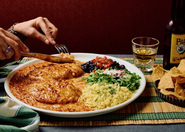 A plate of Mexican food with cheese enchiladas, rice, beans, and salsa, accompanied by chips, a bottle of beer, and a glass of beer on a colorful table mat.