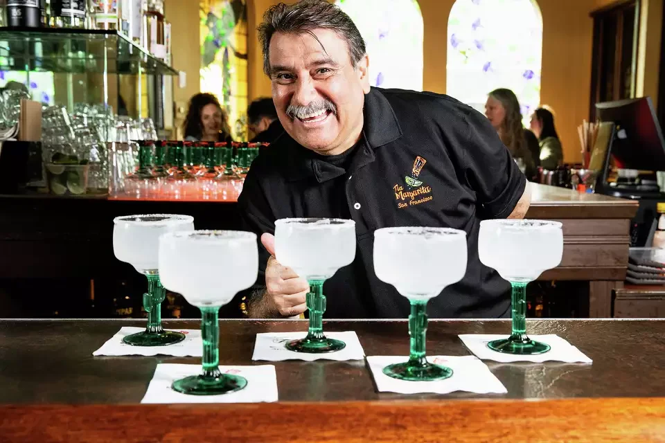 A smiling bartender in a black shirt behind the bar, with six frosted margarita glasses with green stems arranged on the counter, each on a white napkin, inside a lively restaurant or bar.