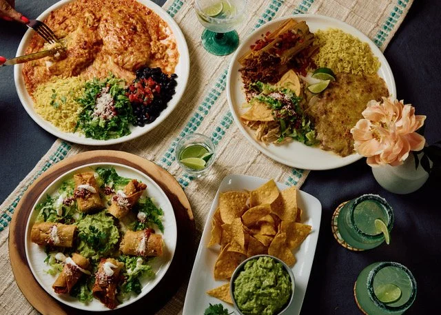 Variety of Mexican dishes including tacos, nachos, rice, beans, and salad on a festive table with drinks and flowers.