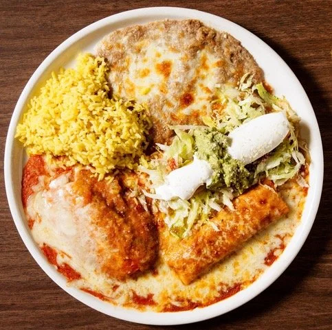 A white plate with Mexican food including a cheese enchilada, a beef enchilada, Mexican rice, refried beans with melted cheese, and a small salad with shredded lettuce, sour cream, and green salsa.