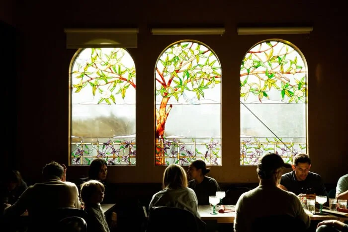 People dining inside a restaurant with stained glass windows depicting a tree with green leaves and white flowers.