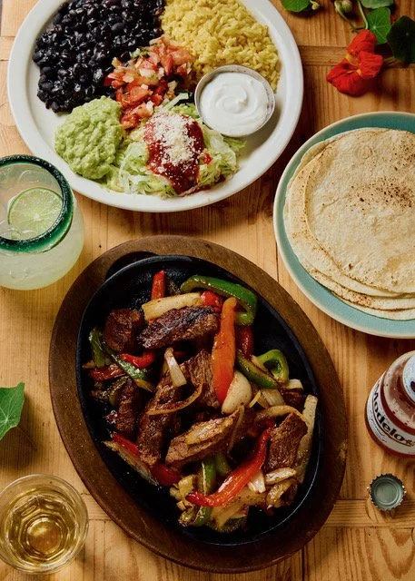 A Mexican meal with a plate of rice, black beans, guacamole, salsa, sour cream, and tortillas, along with a sizzling fajita platter of beef, peppers, and onions, served with drinks and a bottle of hot sauce.