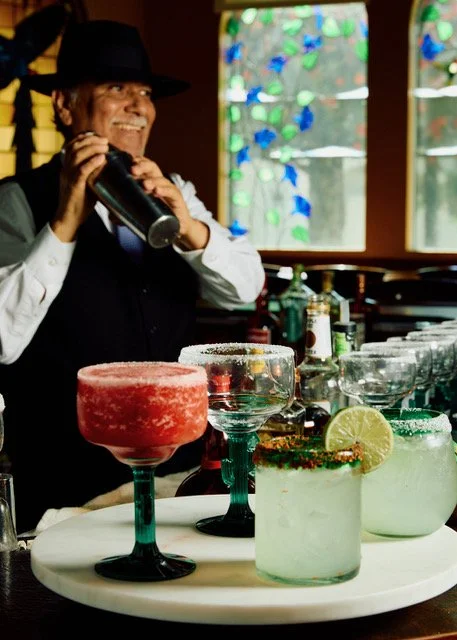A person dressed in black with a white shirt, black vest, and black hat smiling and holding a cocktail shaker in a bar setting with colorful drinks and a stained glass window in the background.