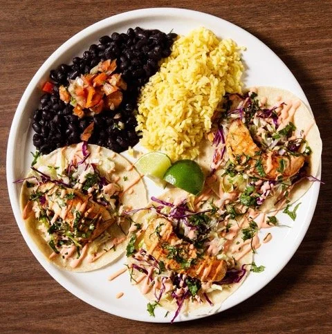 Plate with black beans, yellow rice, grilled chicken tacos with chopped vegetables and lime wedge.