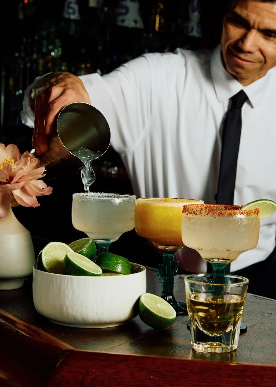 A bartender pouring liquid into a margarita glass, surrounded by lime wedges, a vase with flowers, and other margarita cocktails on a bar counter.