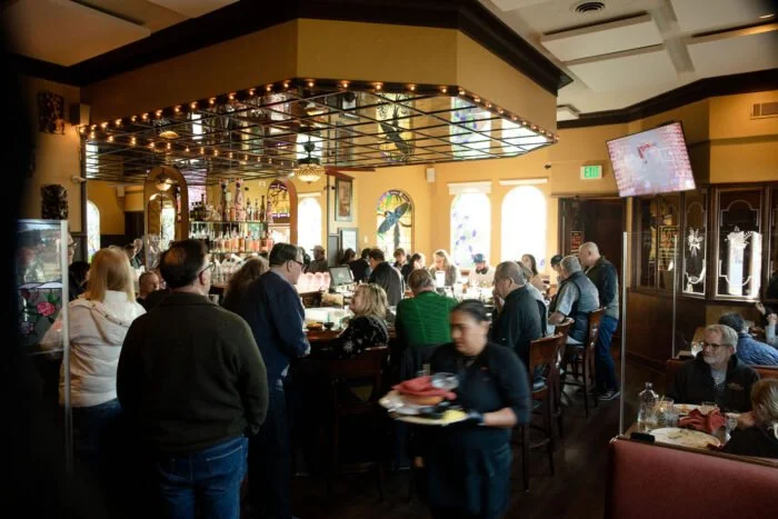 A busy restaurant with people seated at tables and a bar, digital TV screen on the wall, decorative mirrors and stained glass windows, and a server carrying food.