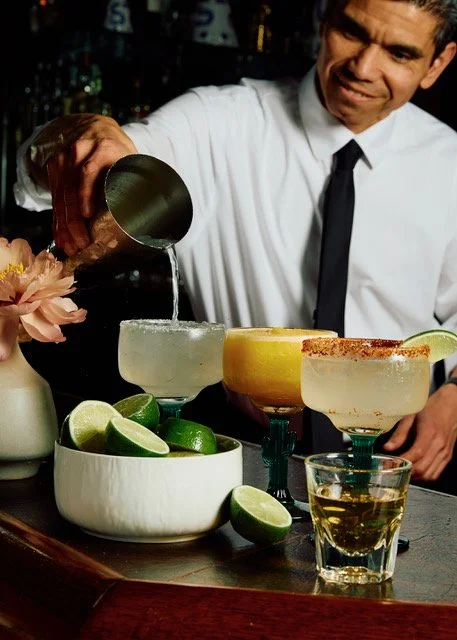 Bartender pouring a clear liquid into a cocktail glass at a bar, with limes and cocktails on the counter.