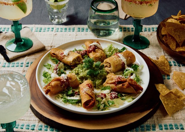 Plate of Mexican taquitos garnished with cheese and cilantro, served with guacamole and salsa, surrounded by drinks and chips.
