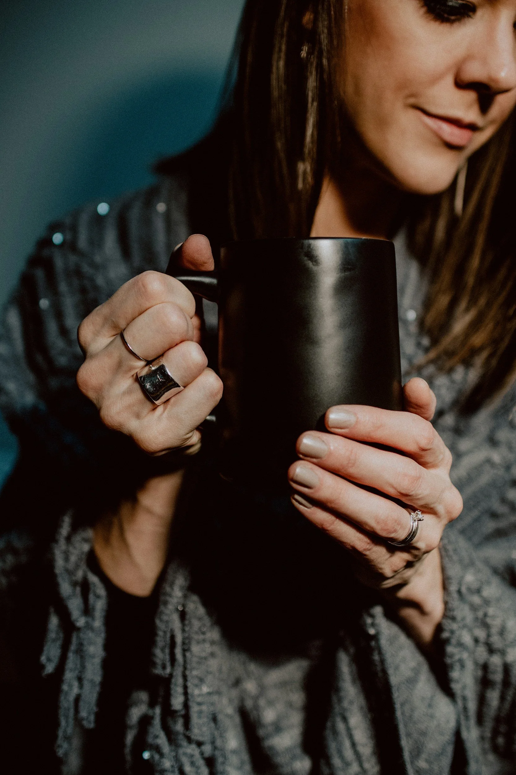 Woman with long brown hair wearing a cozy sweater and holding a black mug, smiling softly.