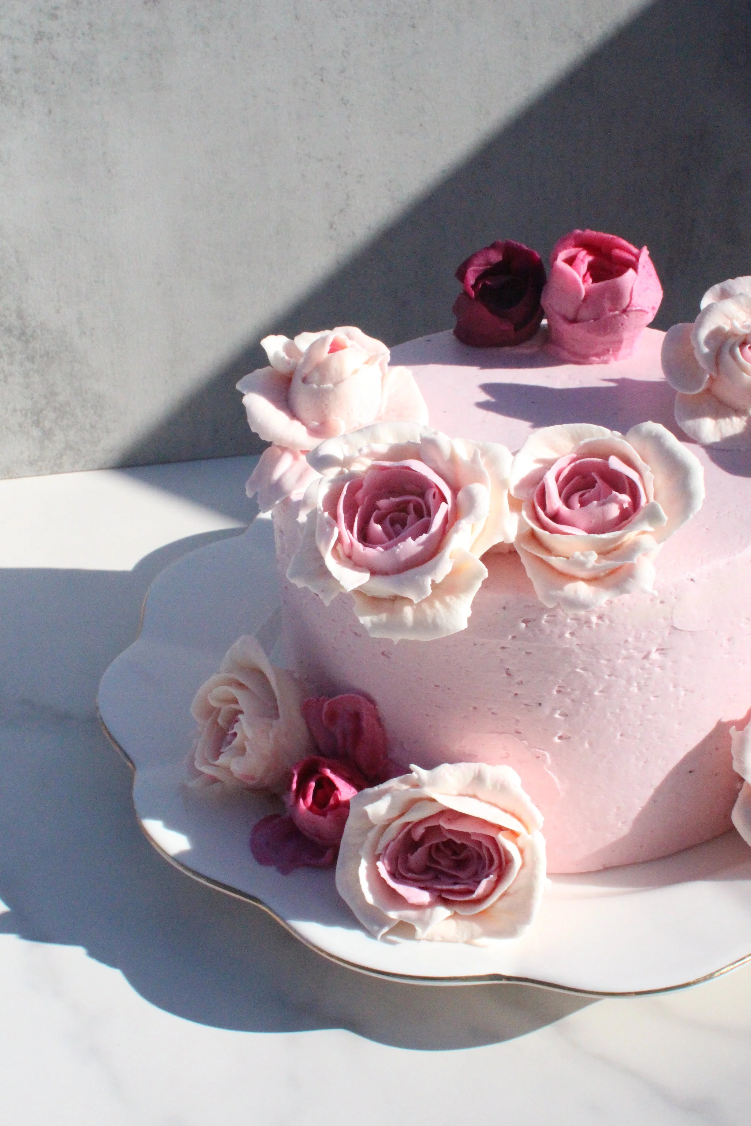 Close-up of a pink cake decorated with pink and white roses made of icing, placed on a white plate with a scalloped gold edge, with shadows cast on a light gray background.