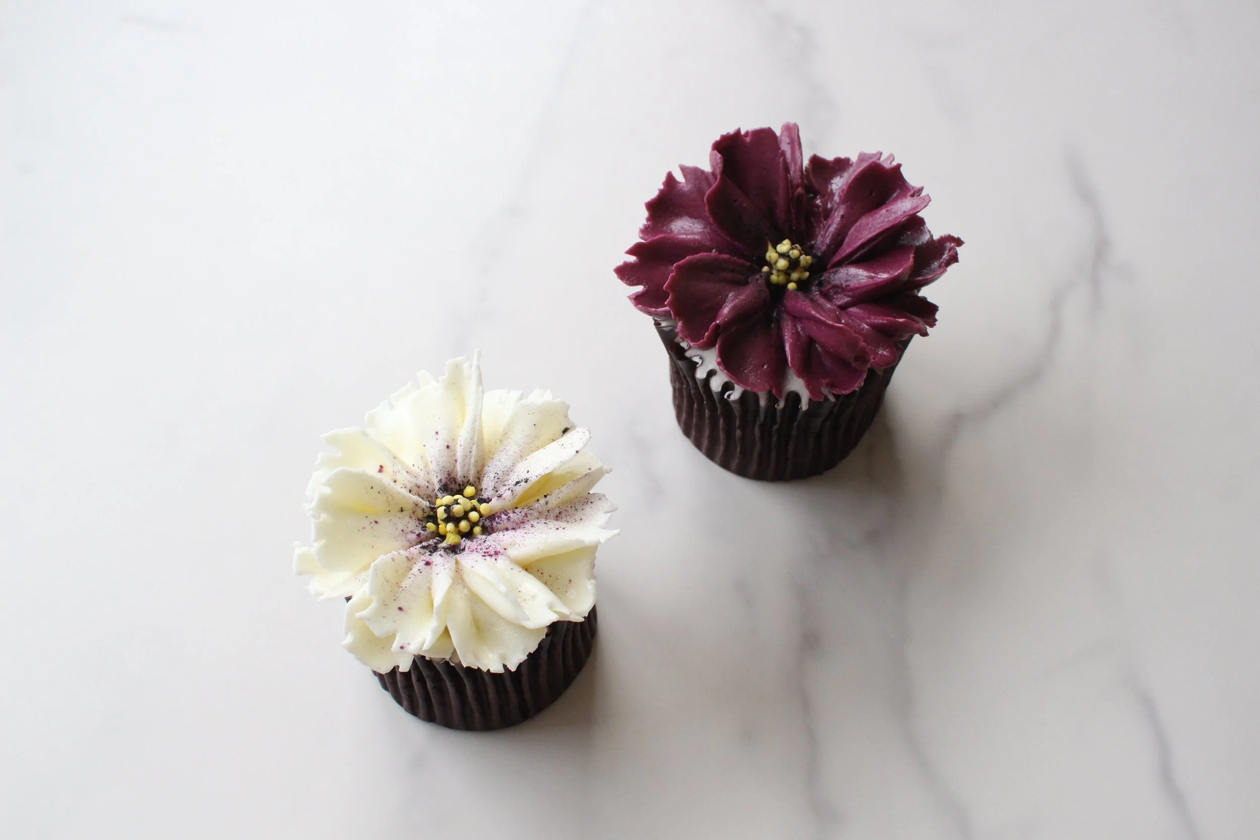 Two decorated cupcakes on a white surface, one with white frosting and purple dust, the other with dark purple frosting resembling a flower, each in a black paper cup.