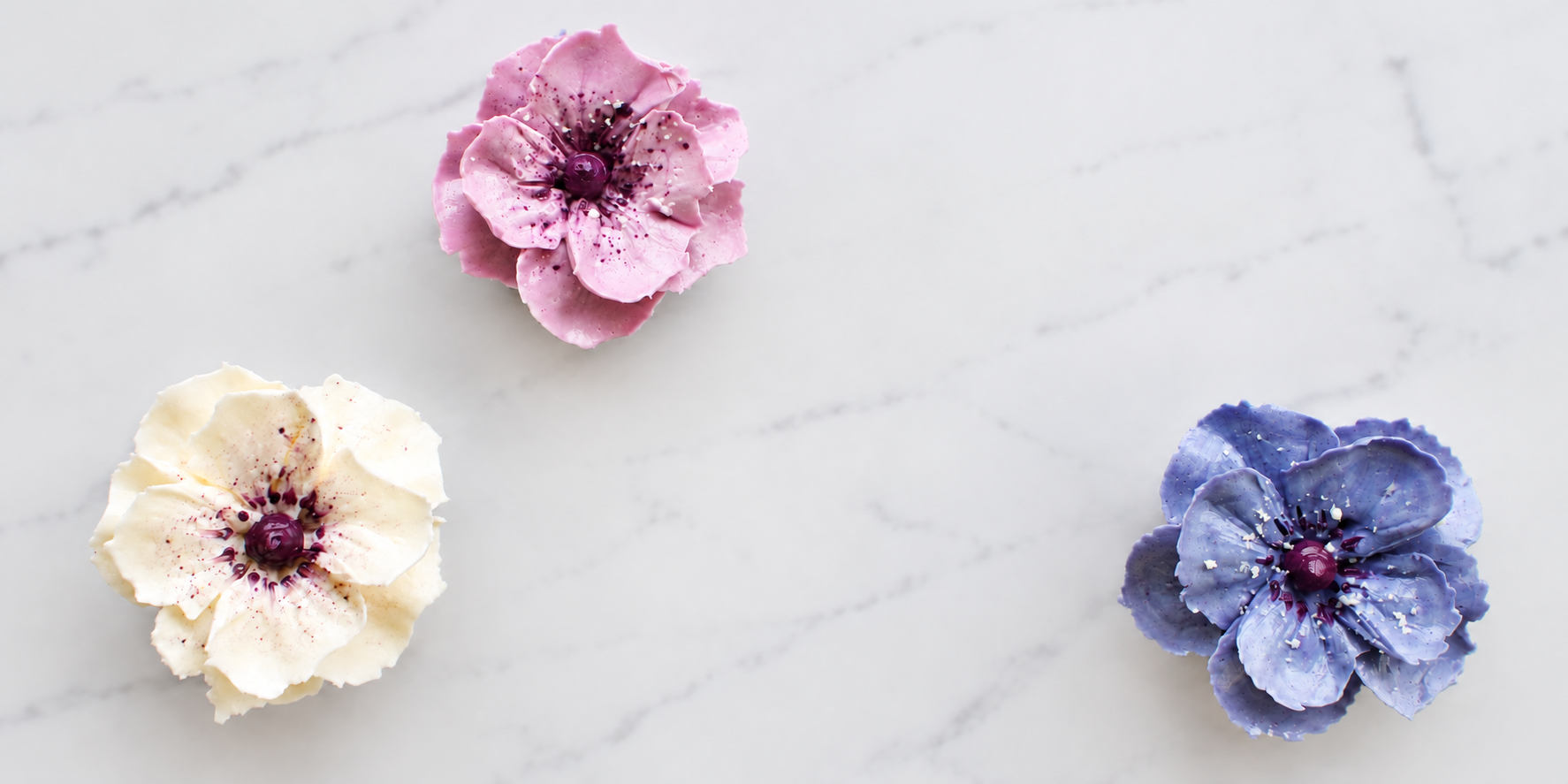 Three decorative flowers in pink, white, and purple, placed on a white marble surface.