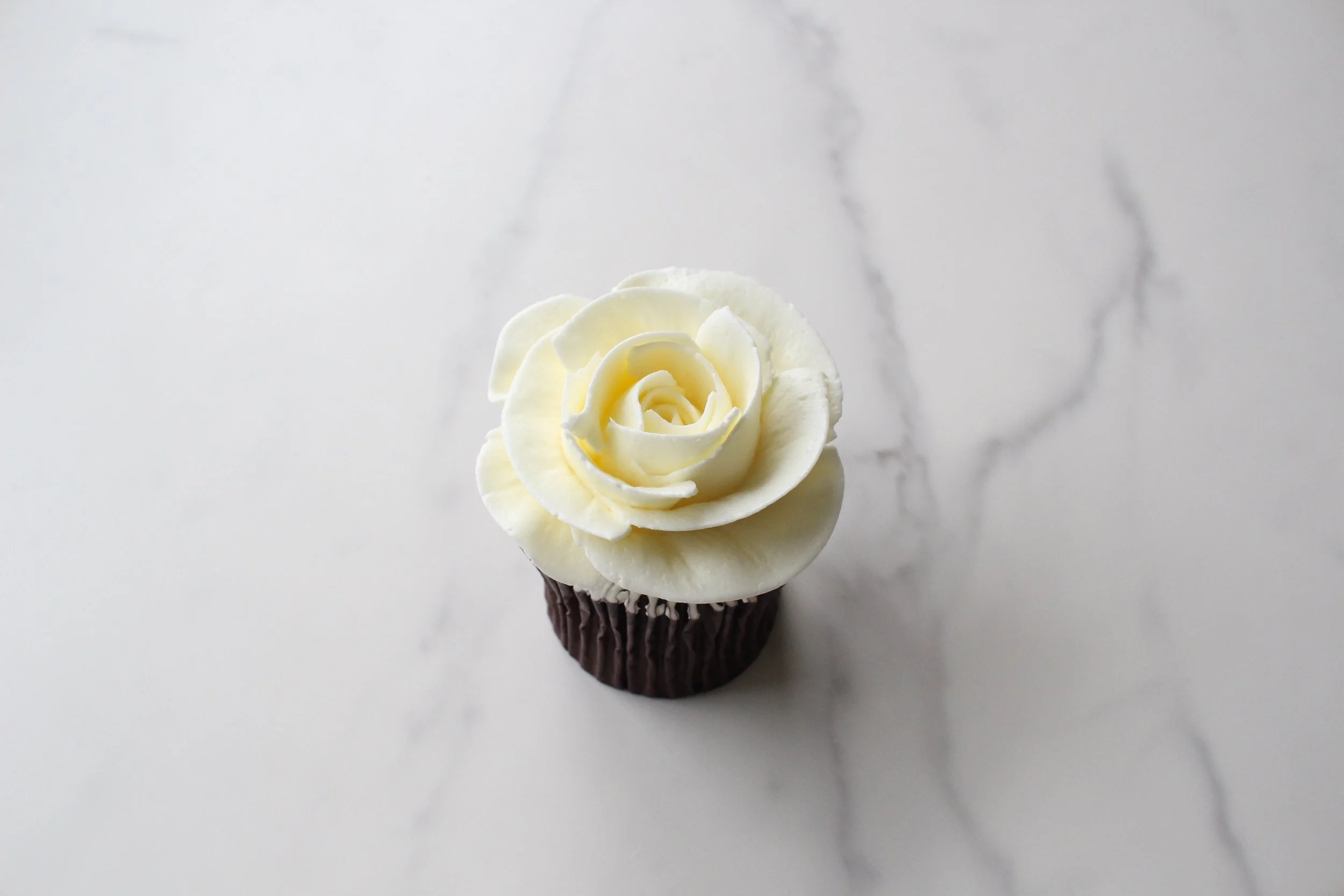 A cupcake with chocolate wrapper topped with white buttercream frosting shaped like a rose, placed on a white marble surface.