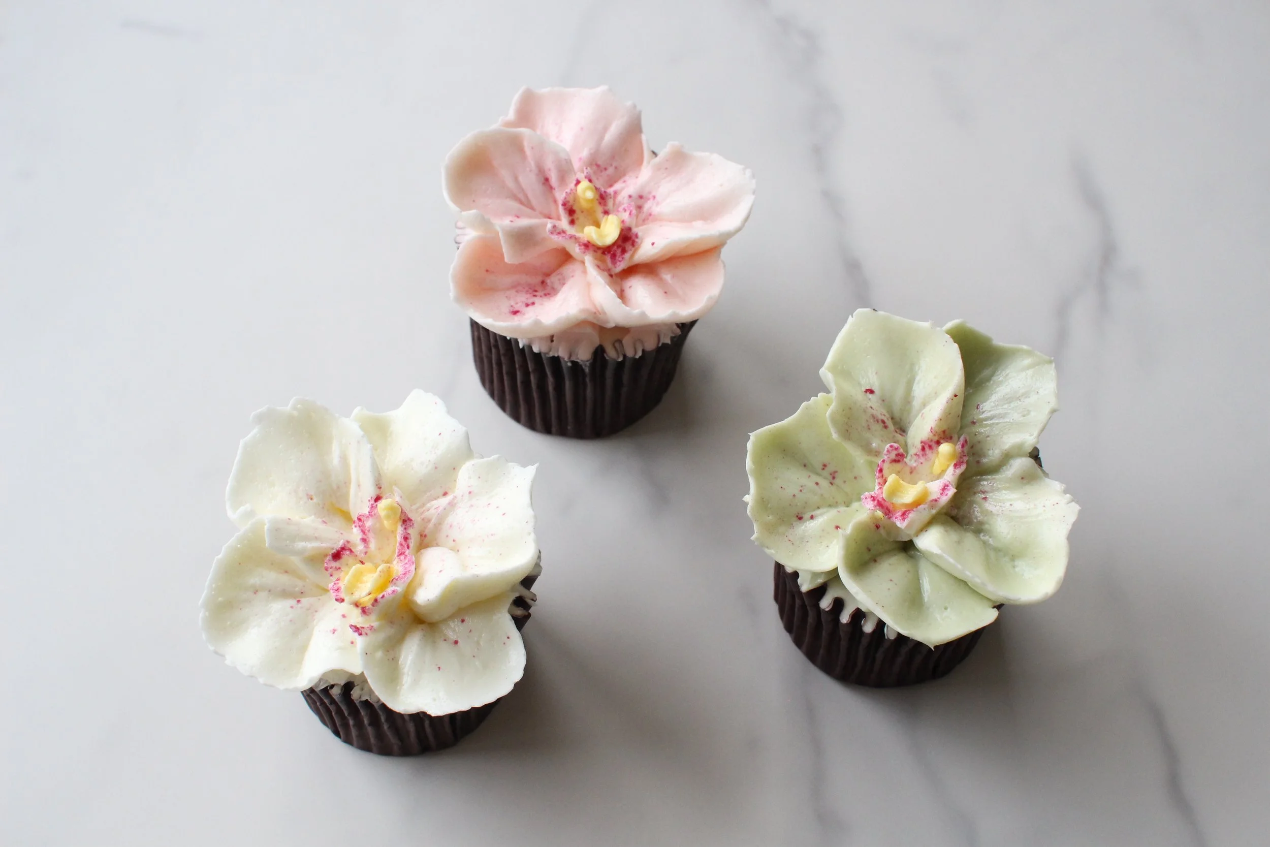 Decorative cupcakes with flower-shaped frosting topped with pink, yellow, and green accents on a white surface.