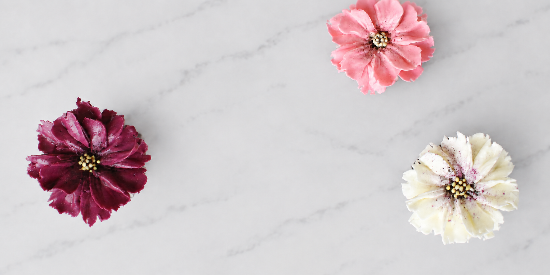 Three decorative flowers on a white marble surface, one pink, one dark pink, and one cream with speckles.