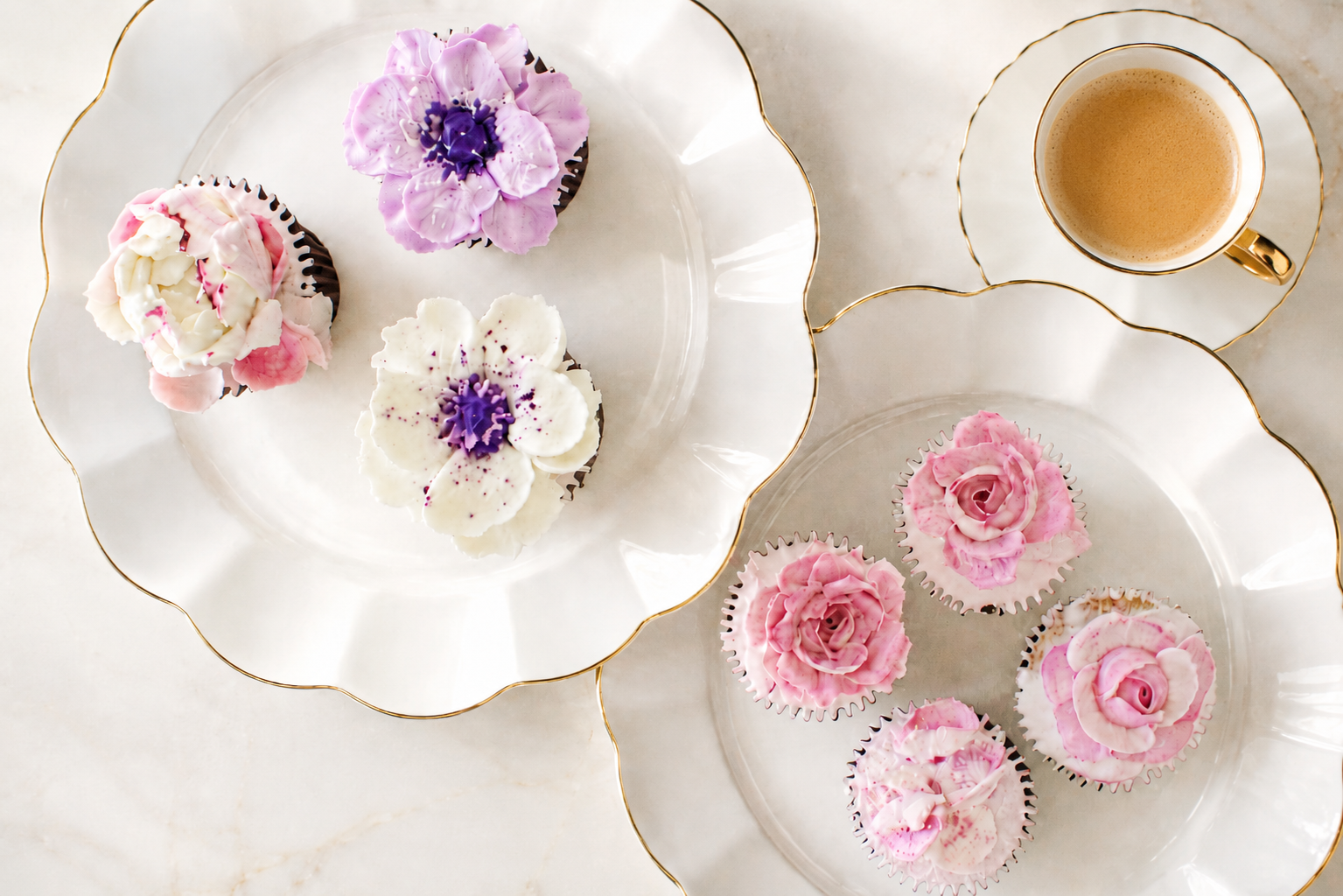 Assorted cupcakes decorated with pink, purple, and white frosting, presented on elegant white plates with gold trim, accompanied by a cup of coffee with cream.