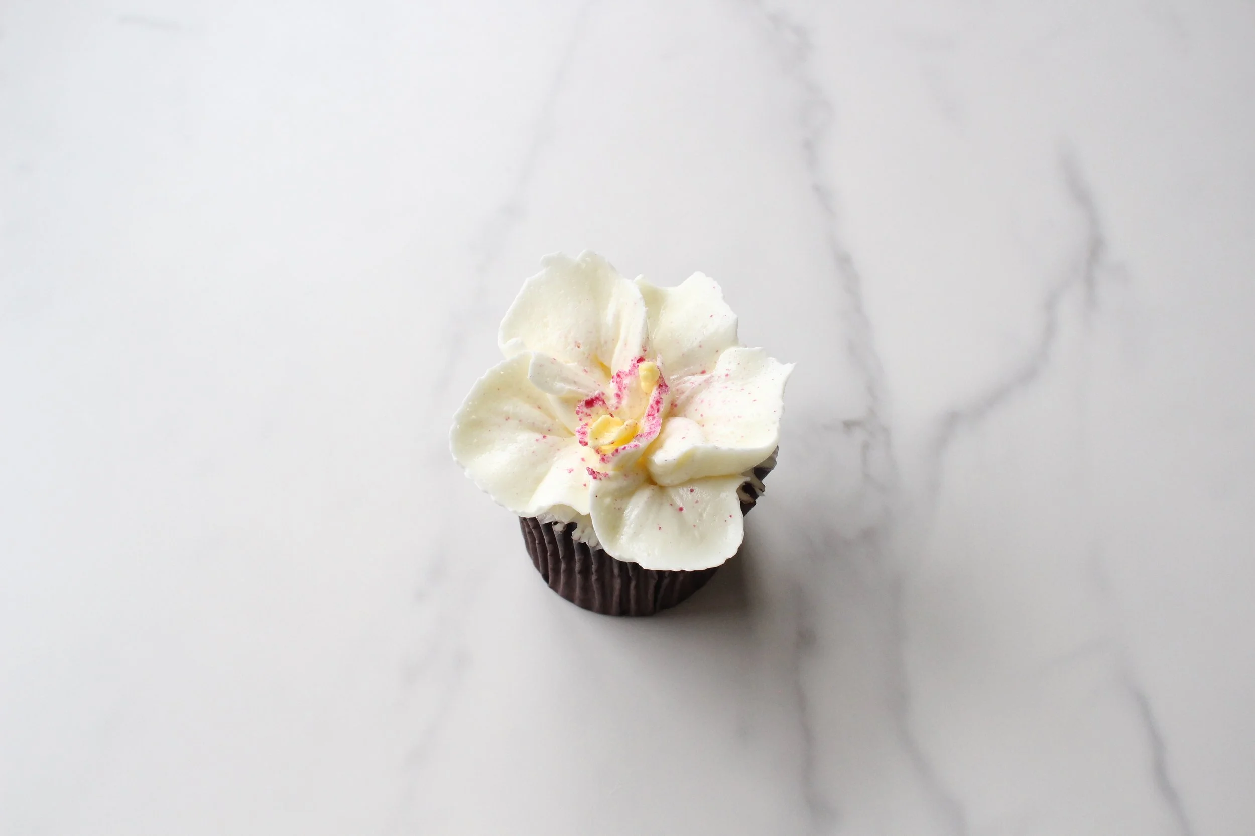 A cupcake with white frosting decorated with pink and yellow sprinkles and a pink flower-shaped candy, in a dark brown cupcake liner, placed on a white marble surface.
