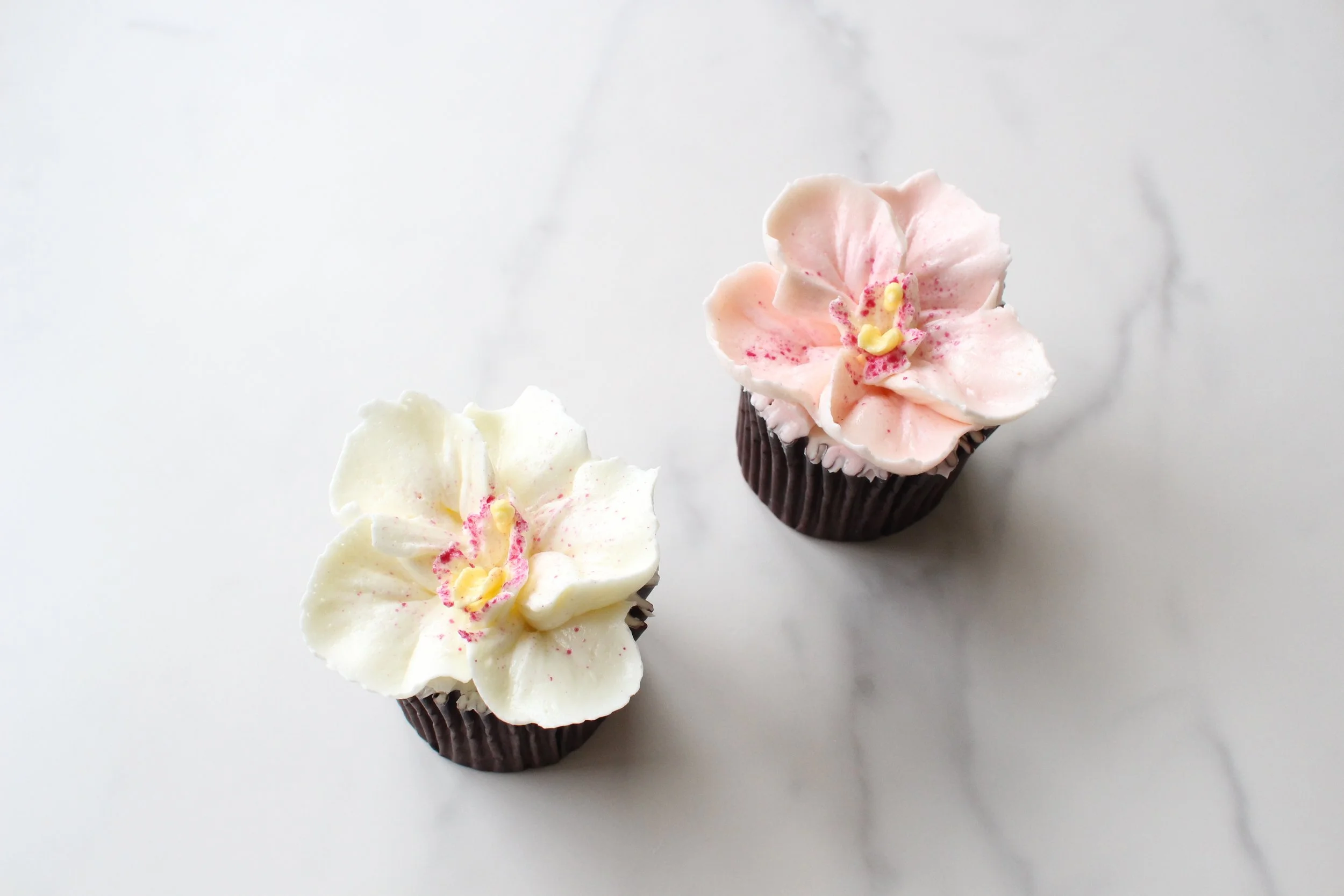 Two cupcakes with flower-shaped frosting in light pink and white, decorated with red sprinkles and small yellow accents, placed on a white surface.