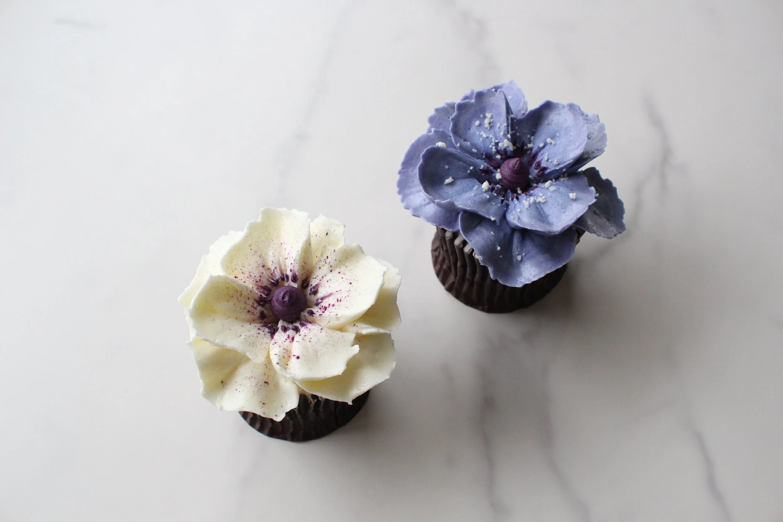 Three cupcakes decorated with flower-shaped frosting in purple, white, and yellow, each topped with a small purple candy and sprinkled with white crumbs, placed on a white surface.