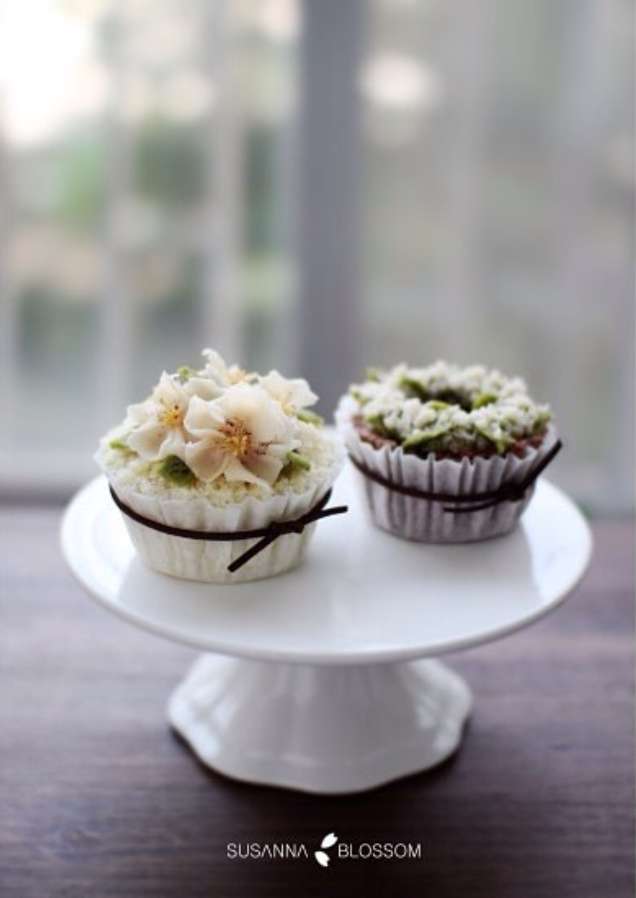 Two cupcakes decorated with floral designs on a white cake stand in front of a window.