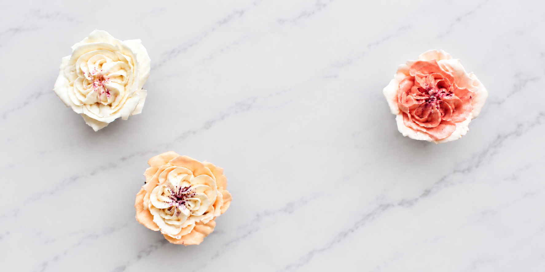 Three cupcakes with floral frosting in white, peach, and pink colors on a white marble surface.