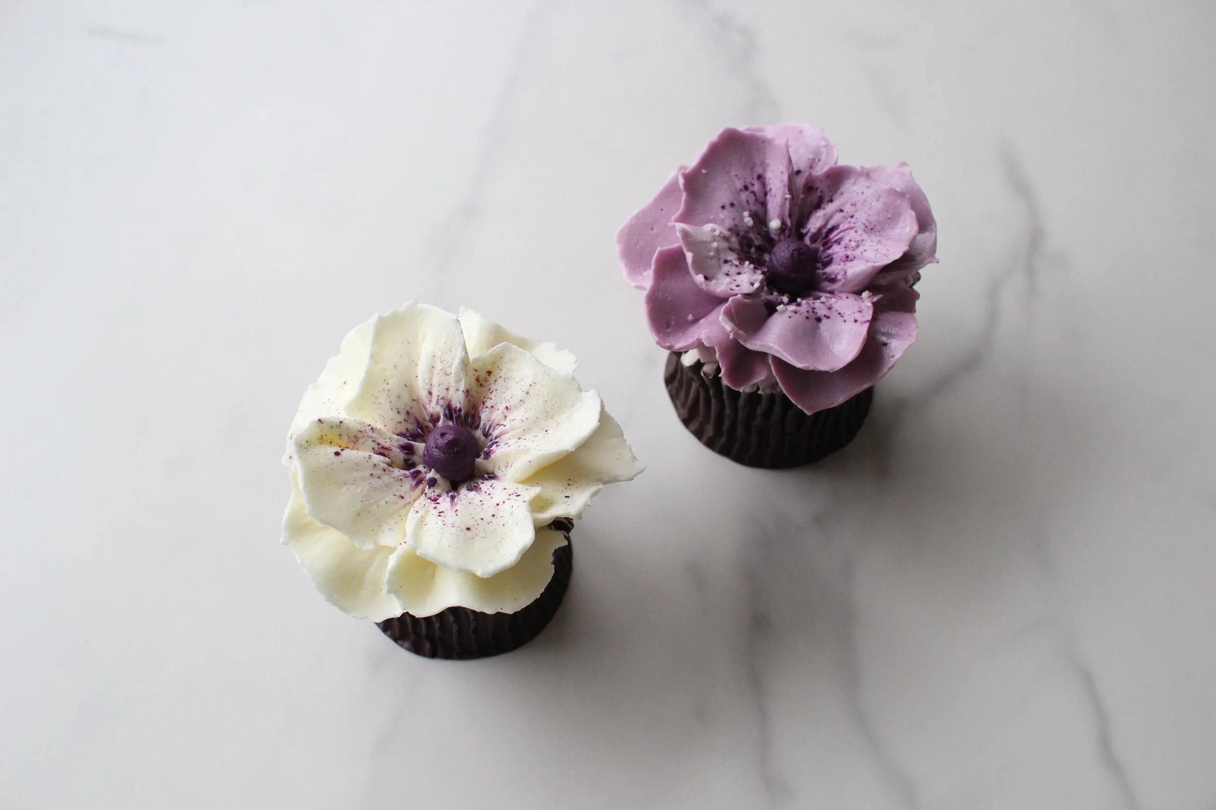 Two cupcakes with flower-shaped frosting, one white with purple details and the other purple with darker purple details, topped with small edible pearls, placed on a white surface.