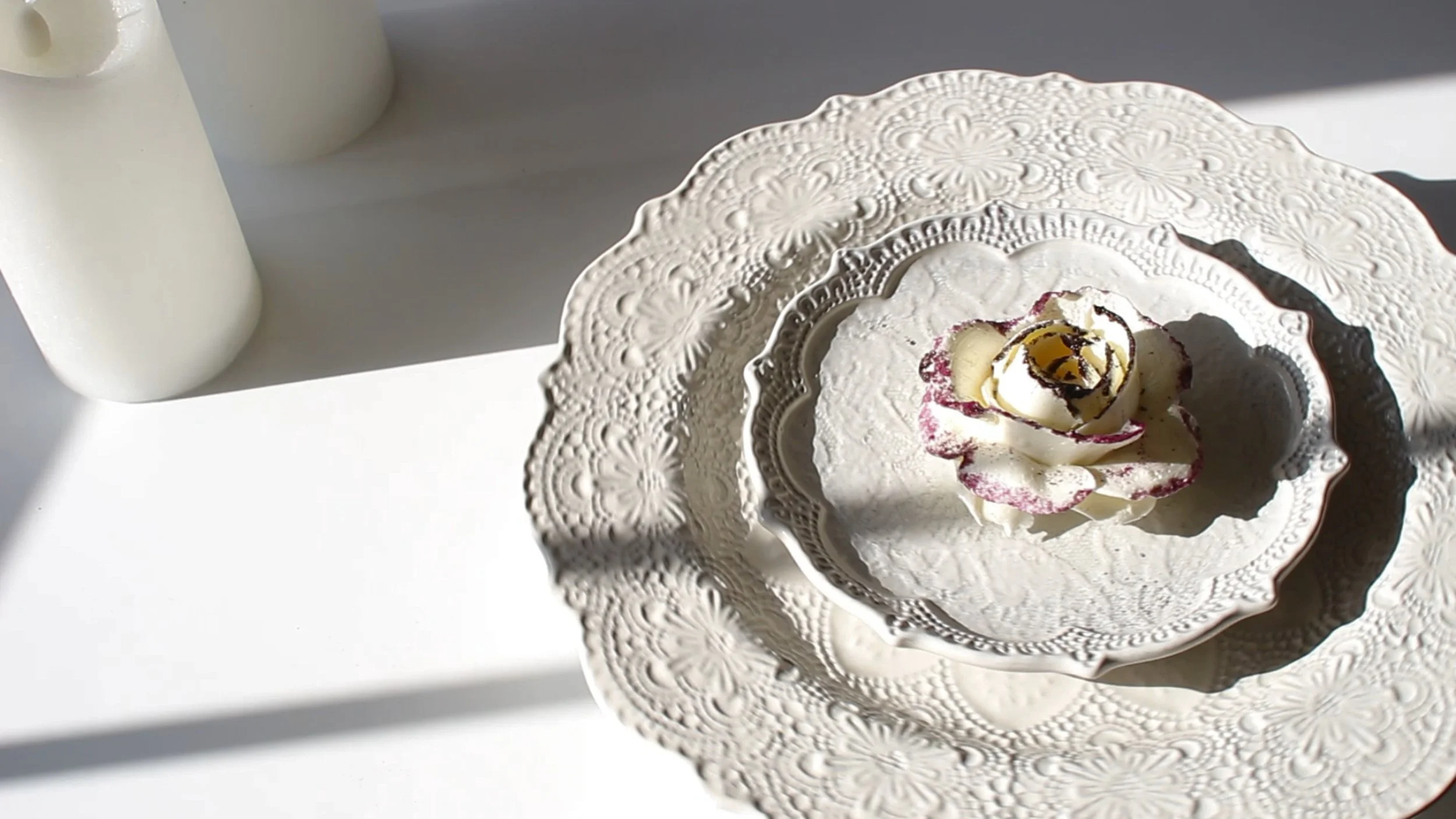 A decorative white ceramic plate with intricate floral patterns, holding a dessert with cream, dark chocolate drizzle, and a rose-shaped decoration, placed on a matching stand.