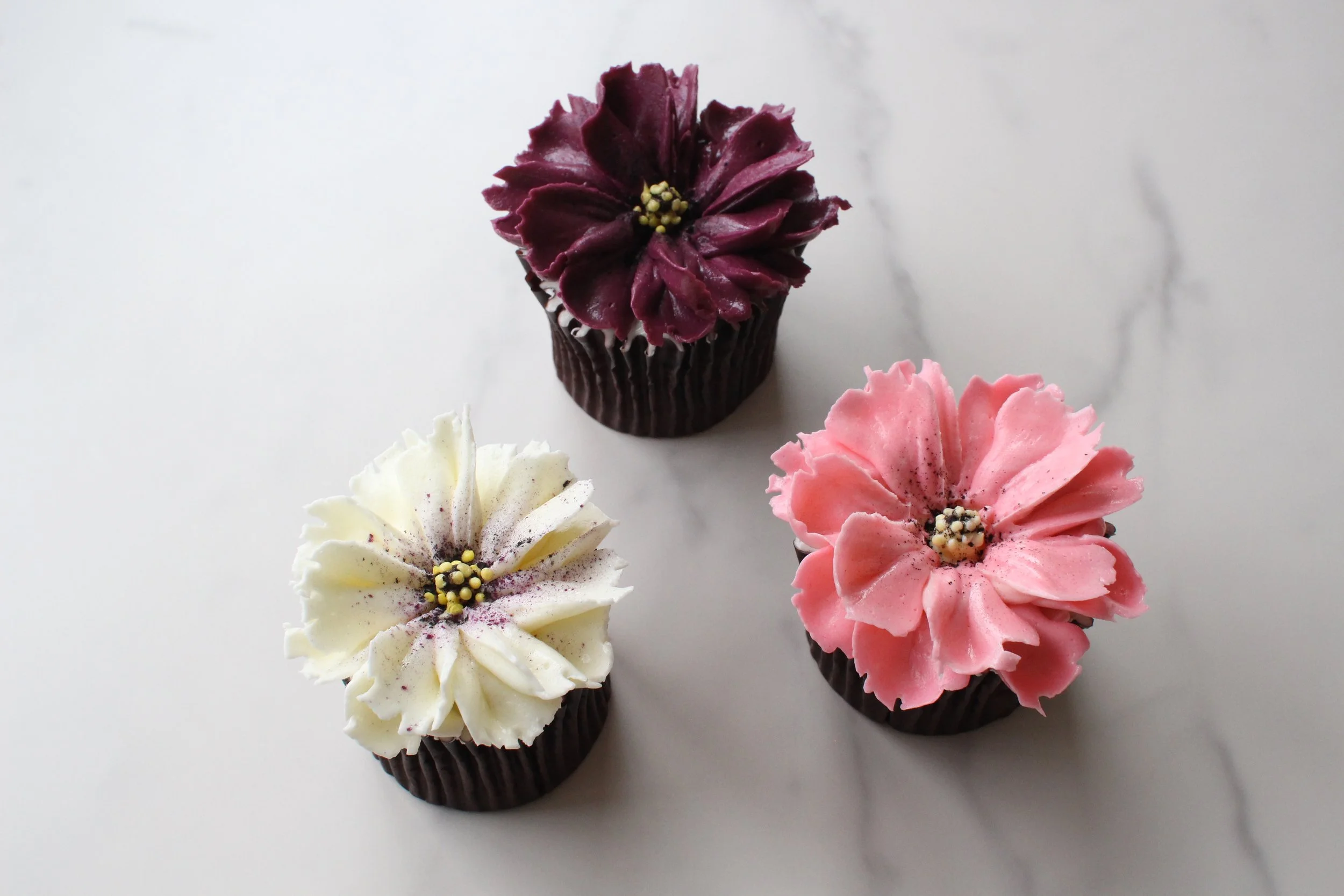 Three cupcakes decorated with flowers made of frosting in white, dark purple, and pink colors, each with a black cupcake liner, placed on a white surface.