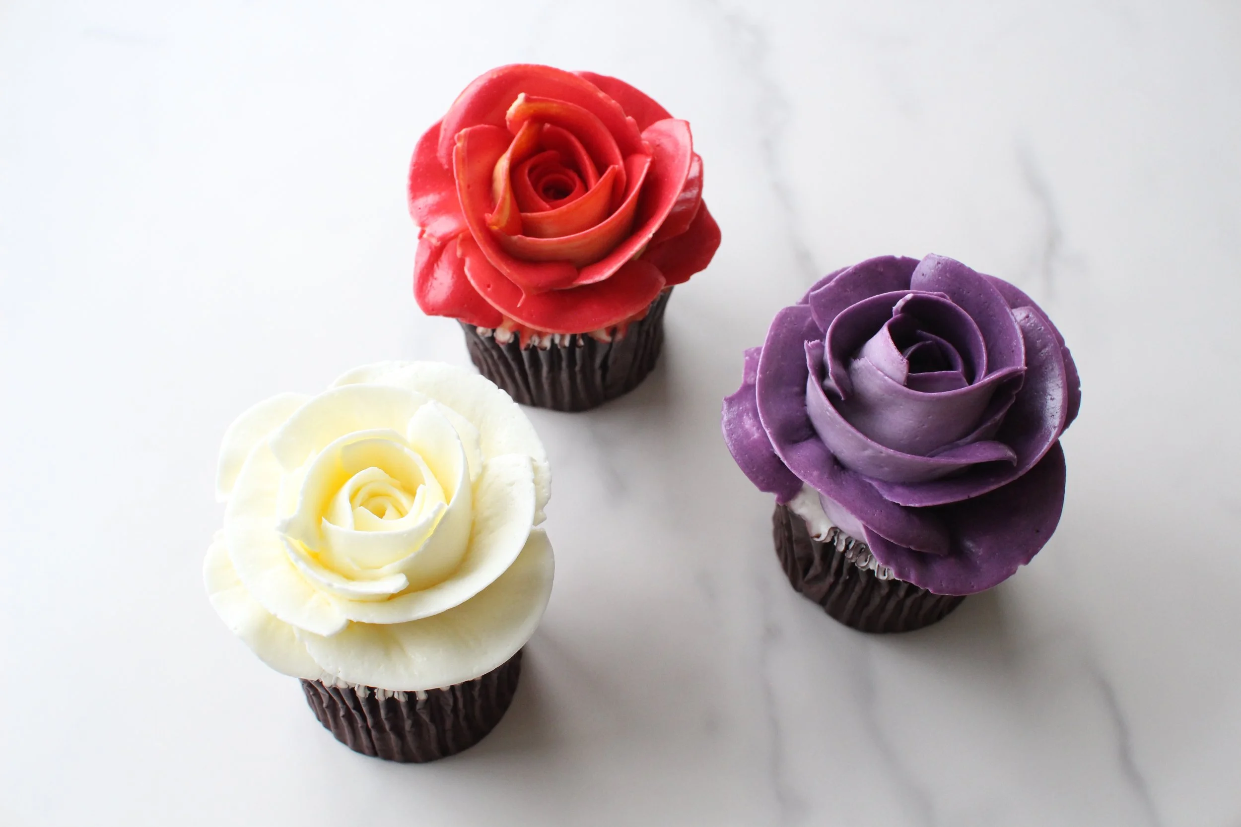 Four cupcakes decorated like roses in red, purple, yellow, and white on a white surface.