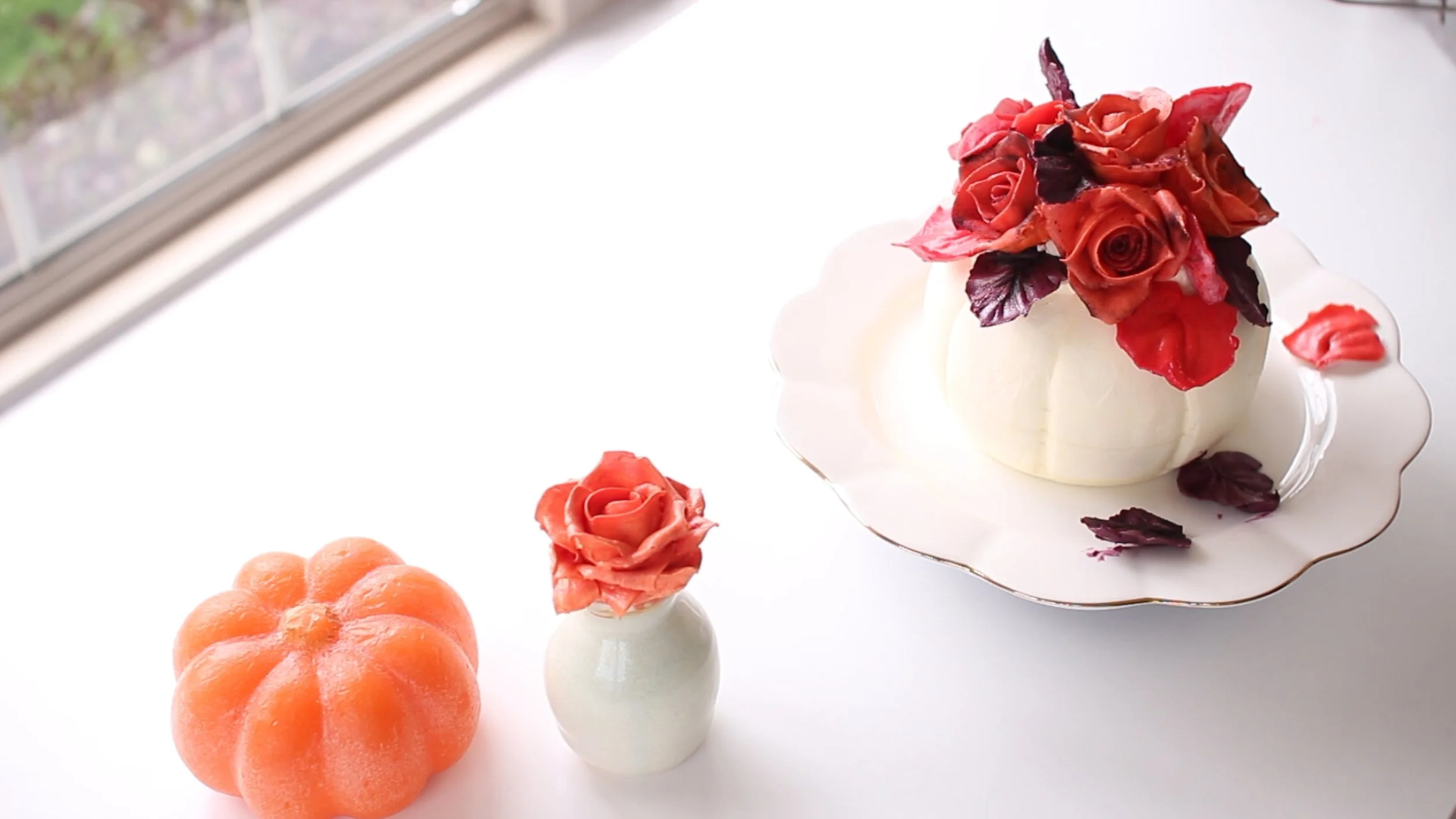 A white dessert decorated with red and purple flower-like shapes on a plate, with small white vases with pink flower shapes and a large orange pumpkin next to it.