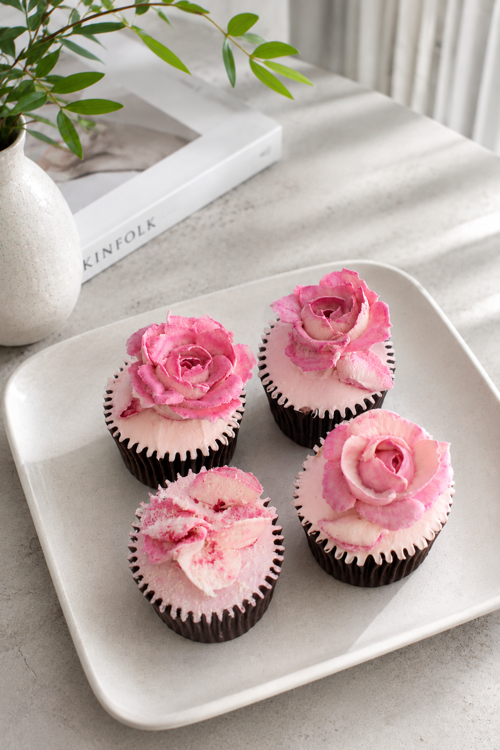 Four cupcakes with pink frosting and rose-shaped decorations on top, arranged on a white square plate.