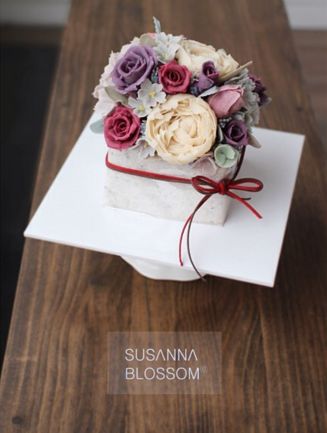 A flower arrangement with various pastel-colored roses and other flowers on a white base, wrapped with a red ribbon, placed on a white cake stand on a wooden surface.