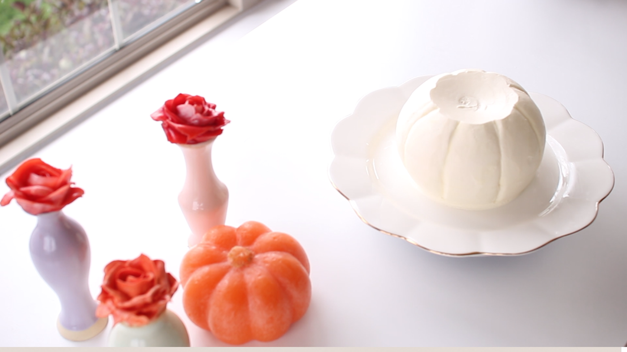 Closed white and gold scalloped edge cake stand with a pumpkin-shaped cake or dessert on top, surrounded by small floral vases with pink and orange roses, and a small orange pumpkin, with a window background.